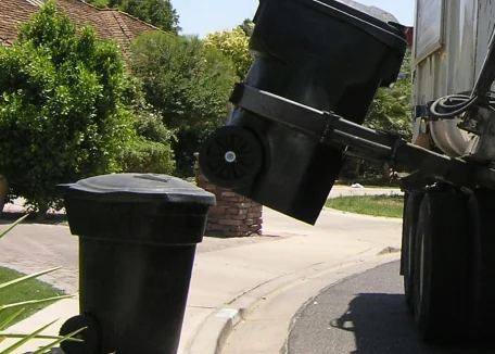 An automated arm on a garbage truck picks up a black garbage bin, another black garbage bin is nearby waiting for pick up