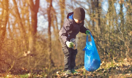 young boy along the edge of a forest holding a blue garbage bag and picking up litter