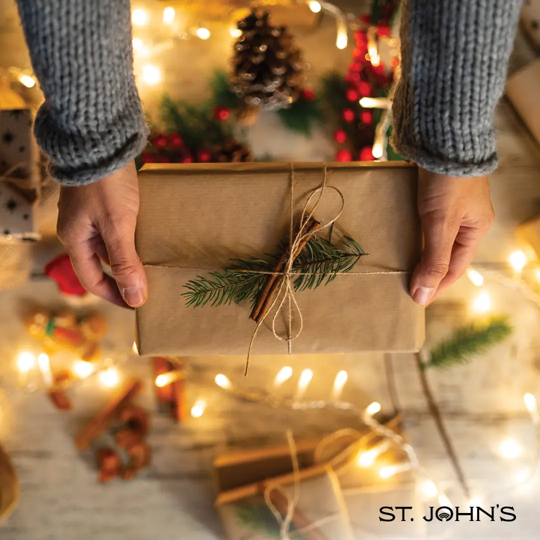hands holding a gift wrapped in brown packing paper and tied with twine and a green conifer tree twig. White decorative lights and green conifer branches are in the background.