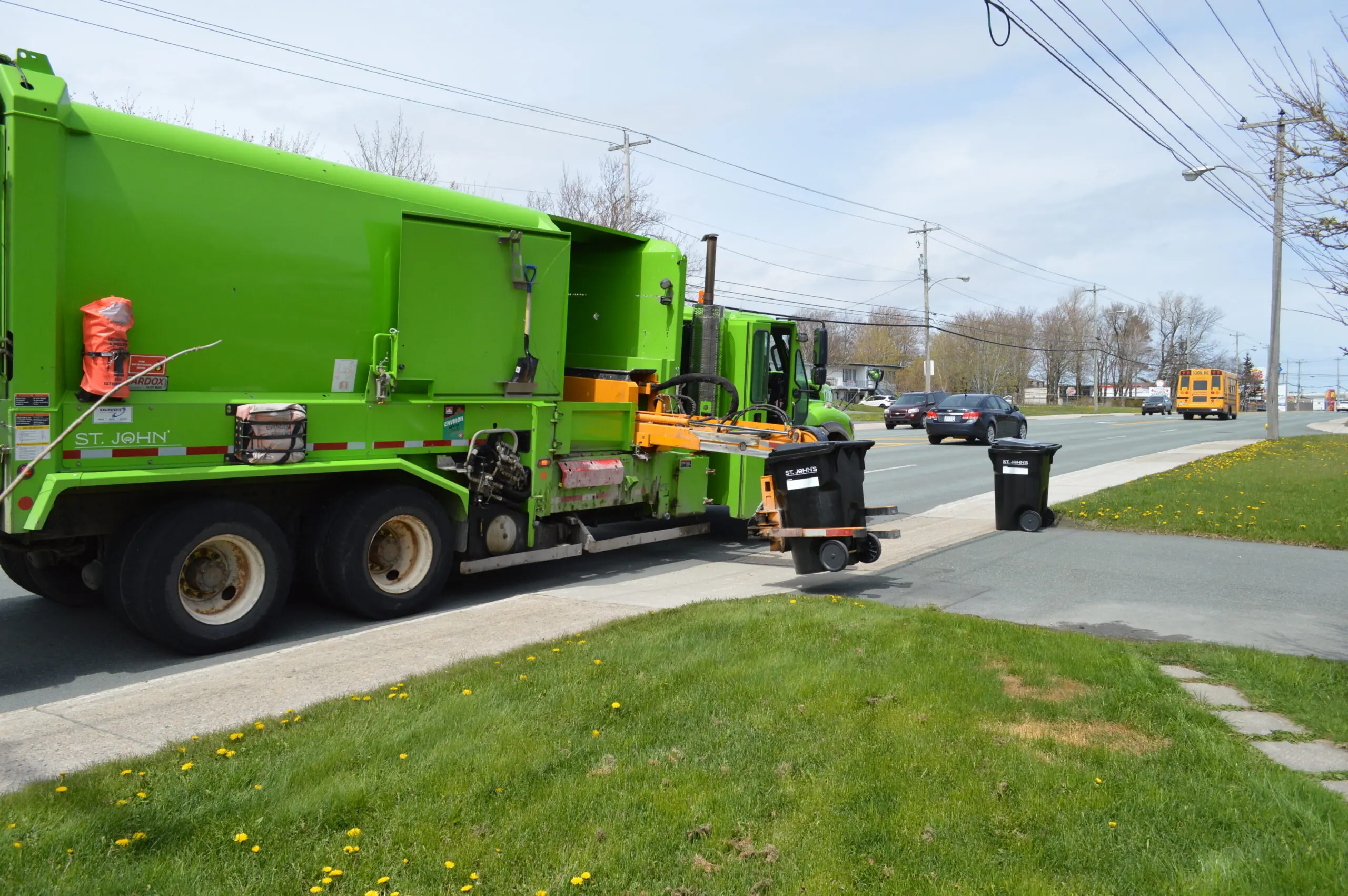 A garbage truck with an automated arm that is picking up a black garbage bin, in the foreground another garbage bin is at the end of the driveway.